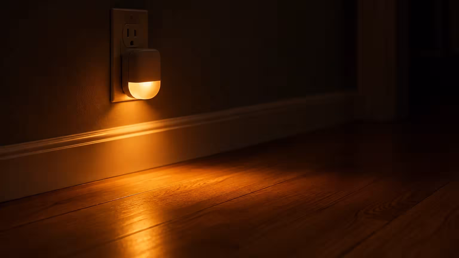 Small plug-in night light glowing warm amber onto a dark baseboard and hardwood floor.