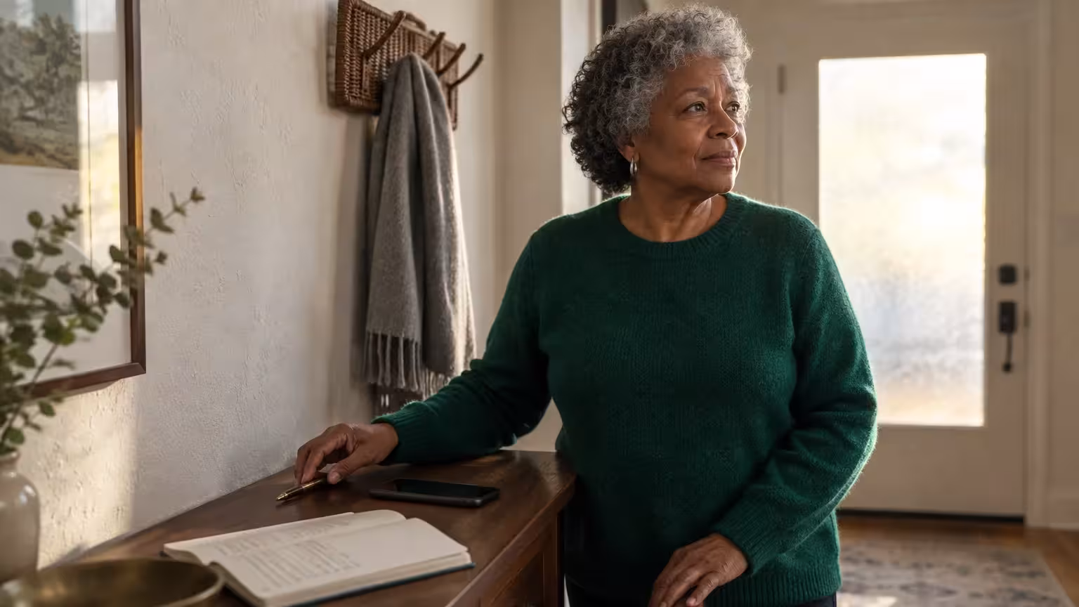 Woman in a green sweater standing by an entryway table with a notebook, pen, and smartphone.