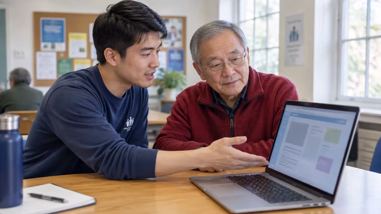 A young man gestures toward a laptop screen while explaining content to an older man wearing glasses.