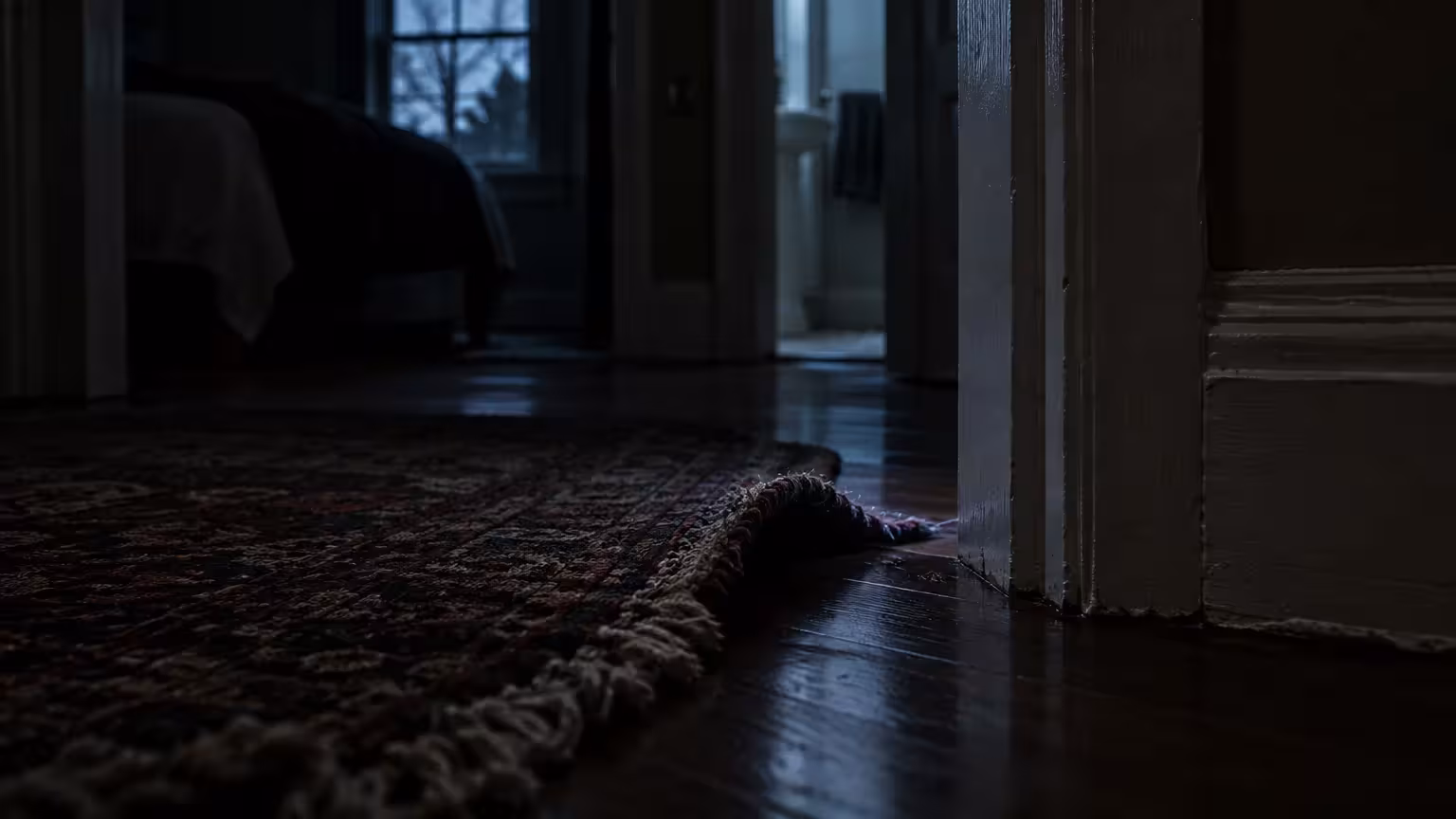 Curled corner of an oriental rug on a dark wood floor in a dimly lit hallway outside a bedroom and bathroom.