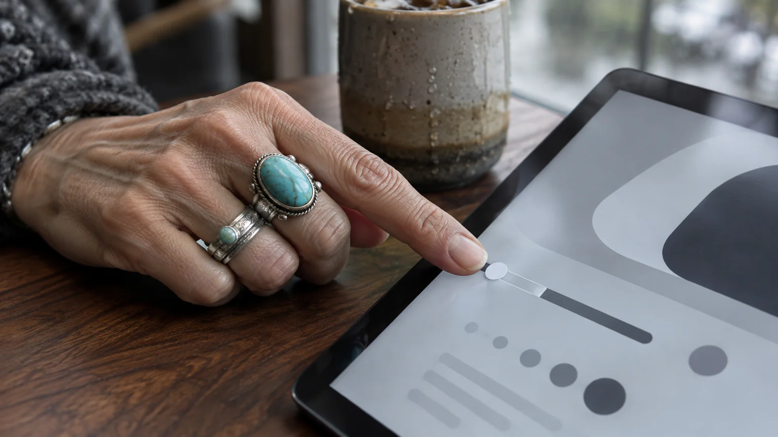 A person wearing turquoise rings adjusts a slider setting on a tablet screen next to a mug on a wooden table.
